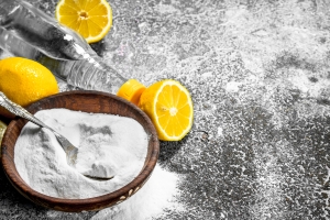 A silver spoon half-buried in baking soda sits on rough stone surface, which has been sprinkled with the same powder, in a wooden bowl. It is framed by bright yellow cut lemons and a bottle of white vinegar.