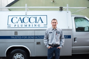 A young man with short hair and a friendly smile stands before a Caccia Plumbing service van. Blue lettering on the side of the white van reads, "Caccia Plumbing Trusted for Generations.