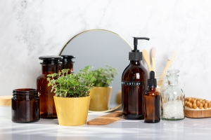 Brown glass bottles and vials sit on a marble countertop beside a small potted plant. A round mirror, a wooden comb, and toothbrushes are arranged neatly beside them.