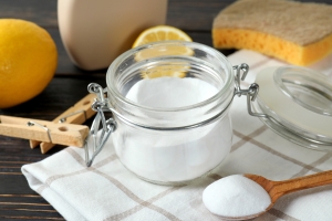 A clear glass jar full of baking soda sits on a gingham cloth next to a wooden spoon, which is also filled with baking soda. Crisp yellow lemons, a yellow and beige sponge, and a wooden spoon containing more baking soda sit next to it.