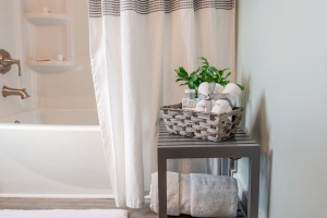 A white molded bathtub with brass fixtures is partially visible behind a crisp white shower curtain. A small gray table holding a woven basket containing rolled towels and a small plant sits in front of it on the floor.