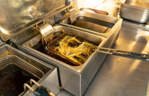 Three silver restaurant deep fryers filled with dark cooking oil sit on an aluminum countertop. The middle one contains a basket filled with french fries, which are actively cooking.