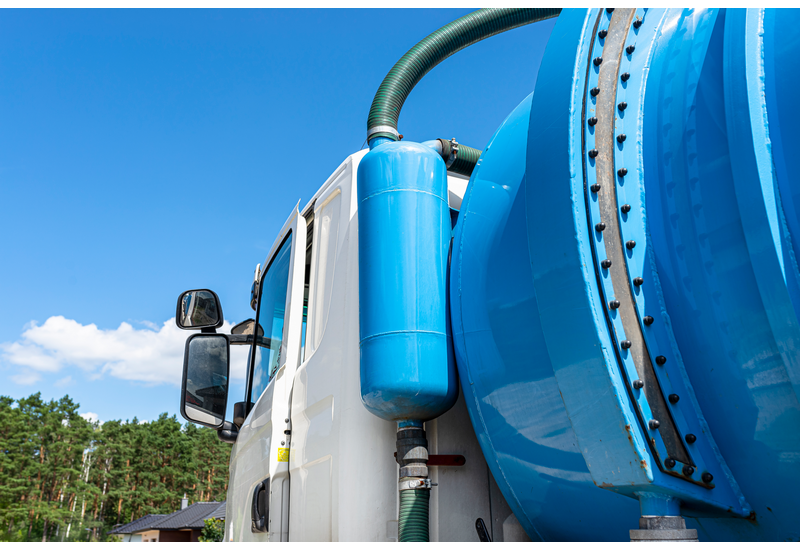 A vacuum truck with specialized equipment for pumping out a septic tank sits parked. The angle of the picture shows the tank, the cab, and a set of green pipes.