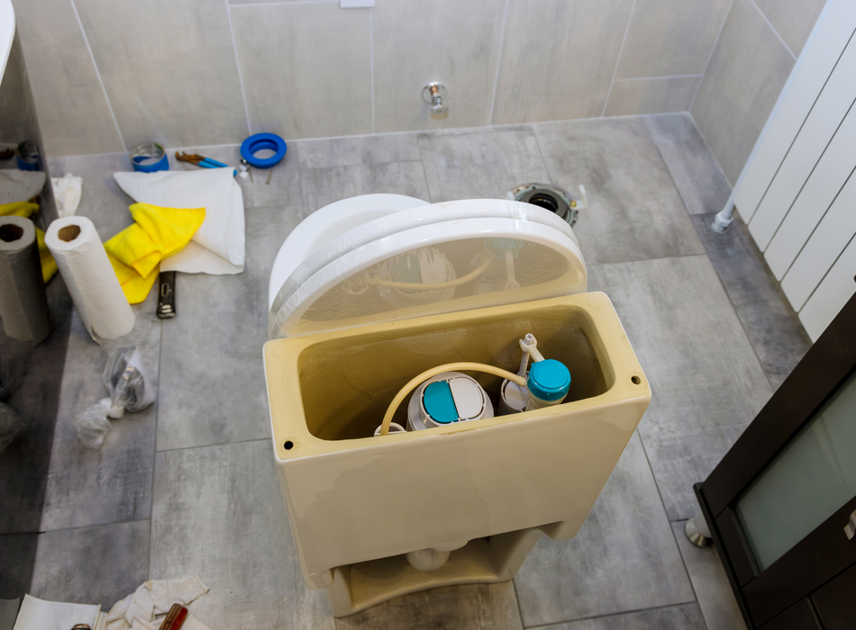 View of the back of a toilet and inner tank, fill valve, and float from above. Various tools and repair items, including a hammer and cloths, lay on the floor nearby. 