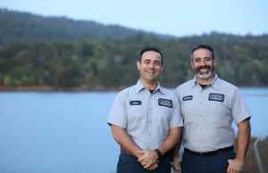 Geno and Anthony Caccia smiling for the camera in front of a body of water and forest backdrop.