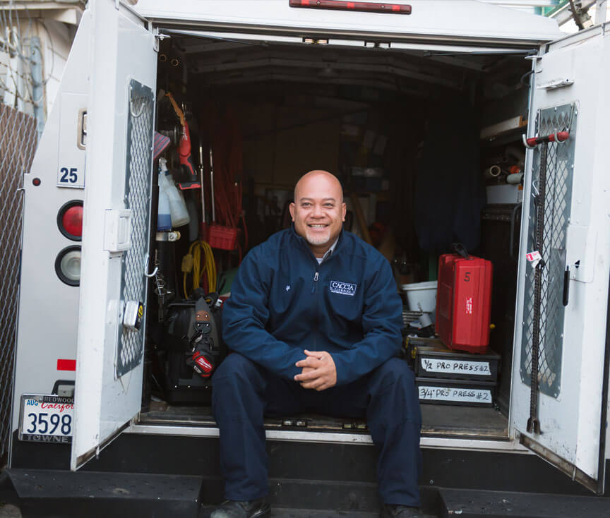Caccia Plumbing plumber sitting in back of an open service van, smiling.